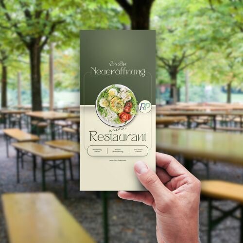Ein DIN Lang Flyer, der in einem Biergarten hochgehalten wird. Es ist Werbung für eine Neueröffnung eines Restaurants.