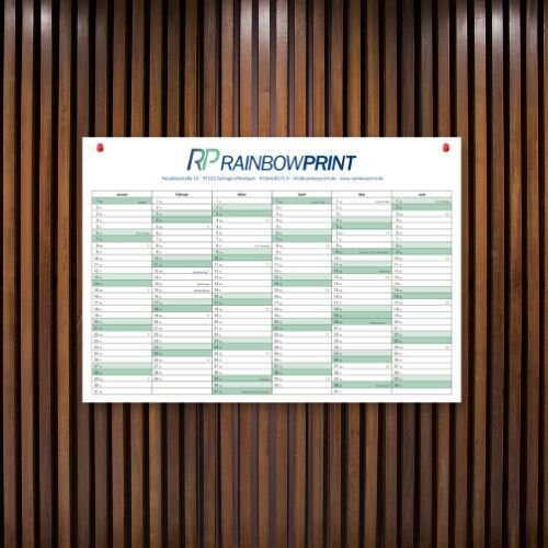 Ein Wandkalender im DIN A Format, welcher an einer hölzernen Akustikwand hängt und mit Stecknadeln befestigt ist.