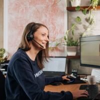 Eine Kundenservice Mitarbeiterin von Rainbowprint, die am Telefon (hier Headset) einem Kunden weiterhilft und dabei vor einem Bildschirm sitzt.